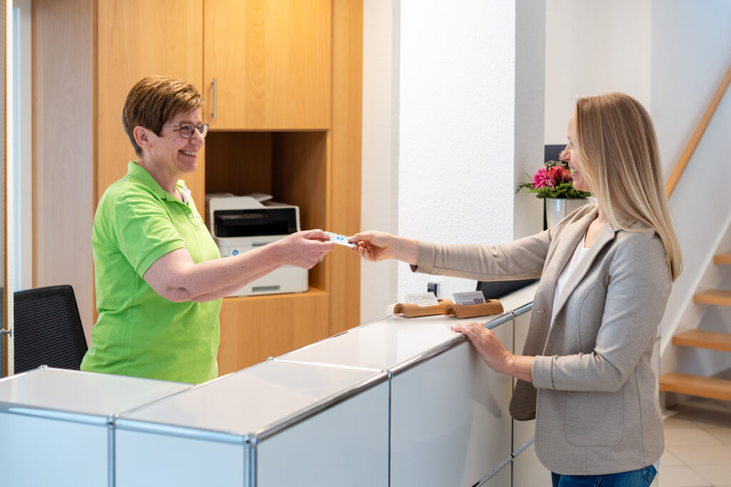 Eine Mitarbeiterin am Empfang gibt einer Kundin eine Visitenkarte der Praxis.