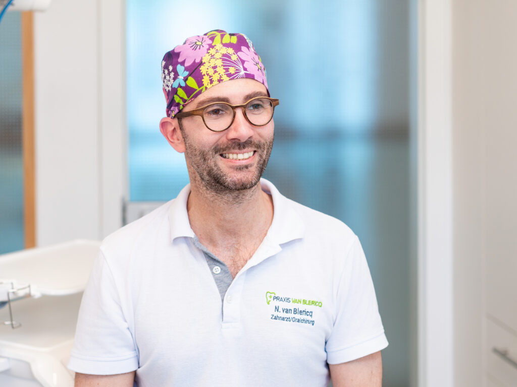 Dr. Nikolai van Blericq sitzt lächelnd mit OP-Haube auf einem Stuhl.